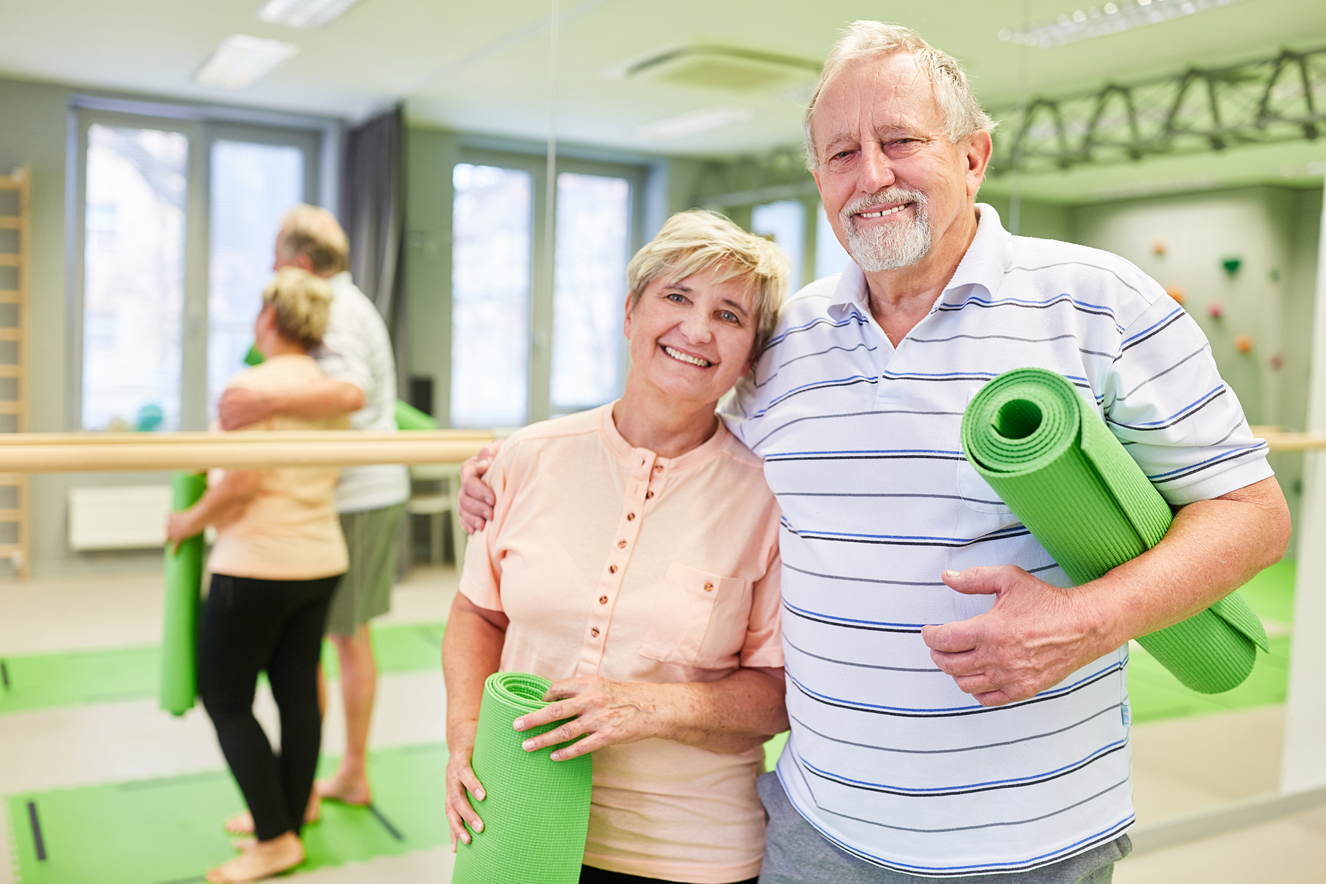 Older adults attending a community yoga or wellbeing class, holding green yoga mats and smiling in a bright fitness studio.